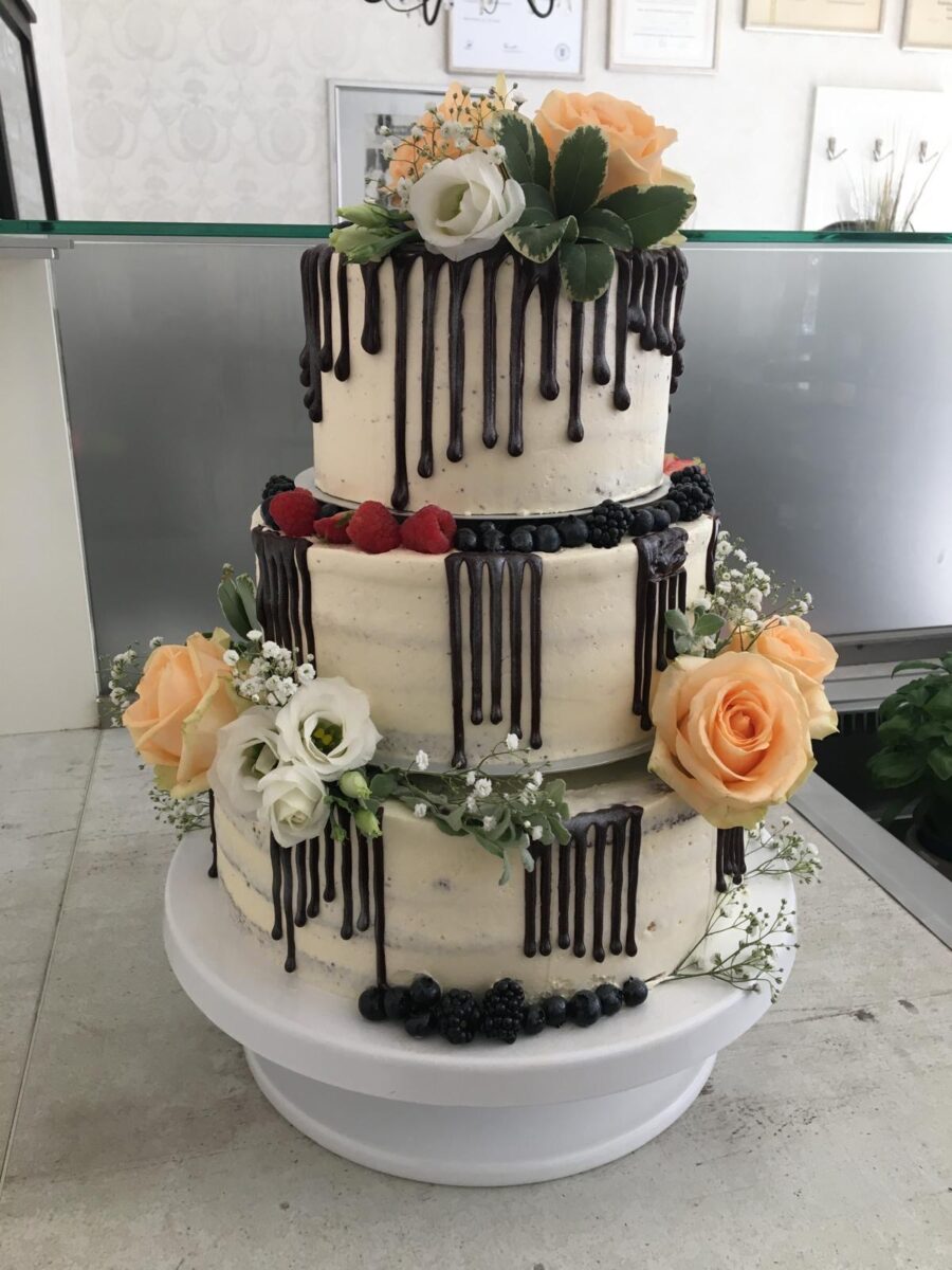 Eine dreistöckige Torte mit weißem Zuckerguss, Schokoladentropfen, frischen Beeren und dekoriert mit pfirsichfarbenen und weißen Rosen, Grünzeug und Babyatemblüten auf einem weißen Tortenständer.