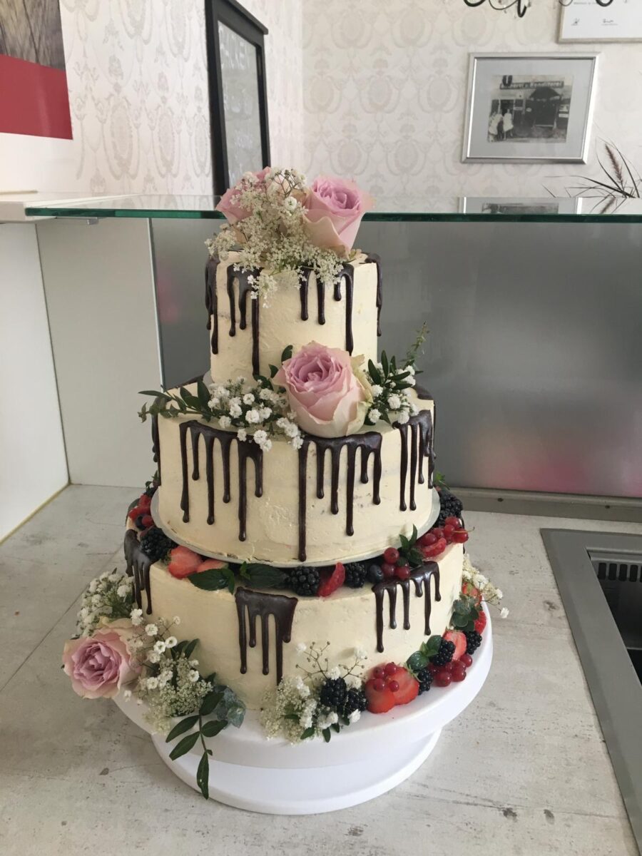 Eine dreistöckige Torte mit cremefarbenem Zuckerguss, Schokoladentropfen, frischen Beeren, rosa Rosen und kleinen weißen Blumen, die auf einem weißen Ständer in einer modernen Küche präsentiert wird.