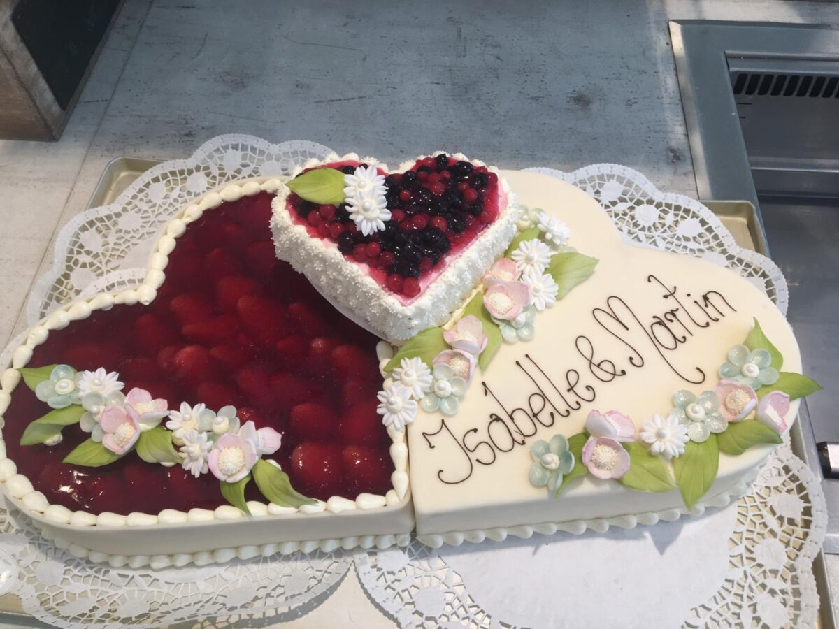 Ein doppelter herzförmiger Kuchen, eine Hälfte mit Erdbeeren, die andere mit weißem Zuckerguss, Blumendekoration und den Worten Isabelle & Martin. Ein kleiner Herzkuchen mit gemischten Beeren liegt obenauf.