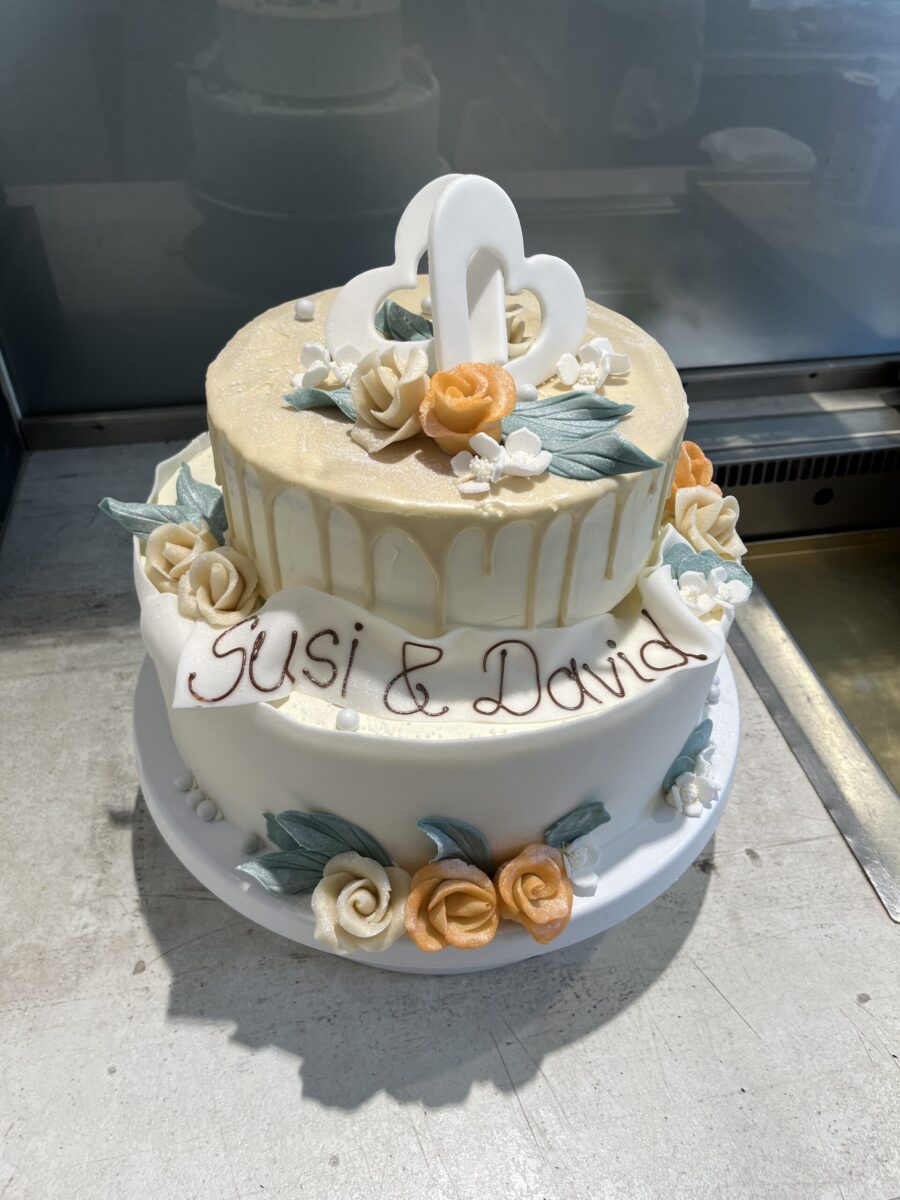 Zweistöckige weiße Hochzeitstorte, verziert mit gelben und weißen Rosen, grünen Blättern und einem weißen Doppelherz als Topper. Auf der unteren Etage steht mit braunem Zuckerguss "Susi & David" geschrieben.