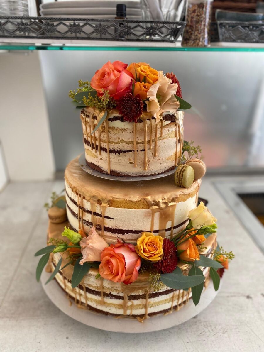Eine dreistöckige nackte Torte mit goldenem Zuckerguss, orangefarbenen und pfirsichfarbenen Rosen, kleinen roten Blumen, Grünzeug und ein paar Macarons auf einem weißen Ständer.