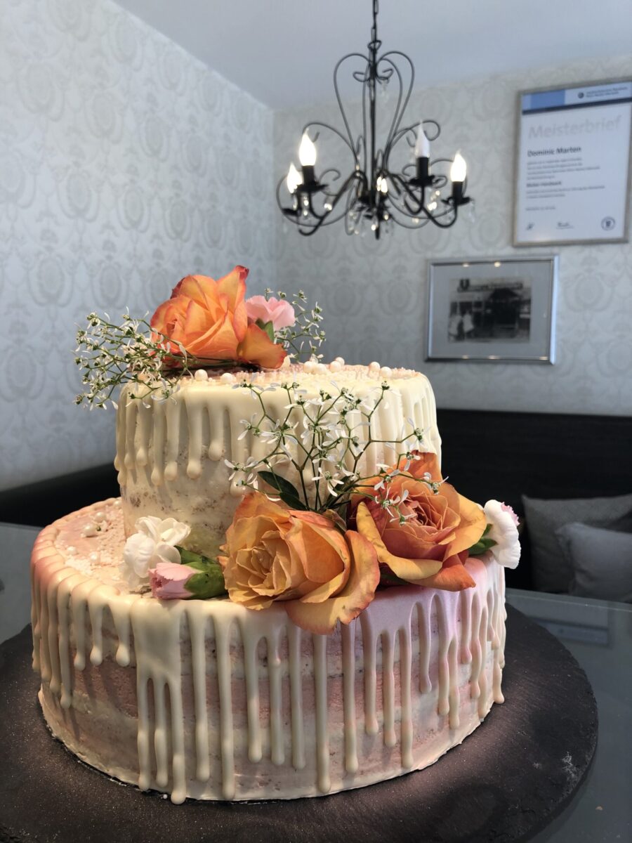 Eine zweistöckige Torte mit weißem Zuckerguss, verziert mit orangefarbenen und pfirsichfarbenen Rosen, Schleierkraut und kleinen weißen Blumen, in einem Raum mit einem Kronleuchter und gerahmten Urkunden an der Wand.