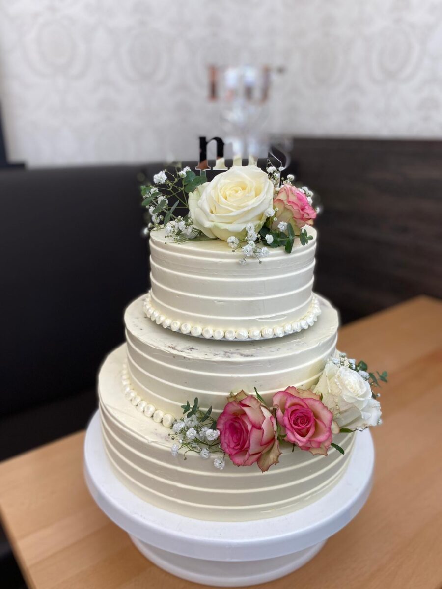 Ein zweistöckiger weißer Kuchen, verziert mit perlenartigem Zuckerguss und gekrönt mit frischen weißen und rosafarbenen Rosen, Schleierkraut und grünen Blättern, auf einem weißen Kuchenständer auf einem Holztisch.