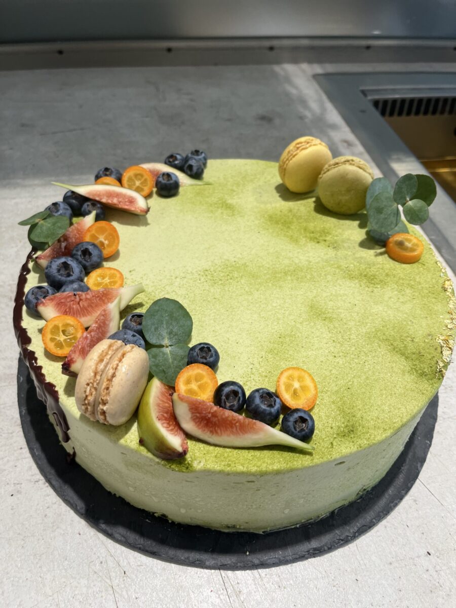 Ein runder Matcha-Kuchen, belegt mit Macarons, geschnittenen Feigen, Blaubeeren, Kapstachelbeeren und Eukalyptusblättern, dekorativ auf einer Metalloberfläche angeordnet.