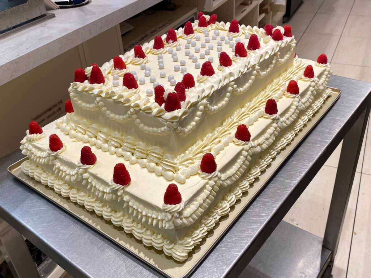 Eine große rechteckige, zweistöckige Torte mit weißer Schlagsahne, roten Himbeeren und kleinen weißen Bonbonkugeln steht auf einem Tablett in einer Küche.