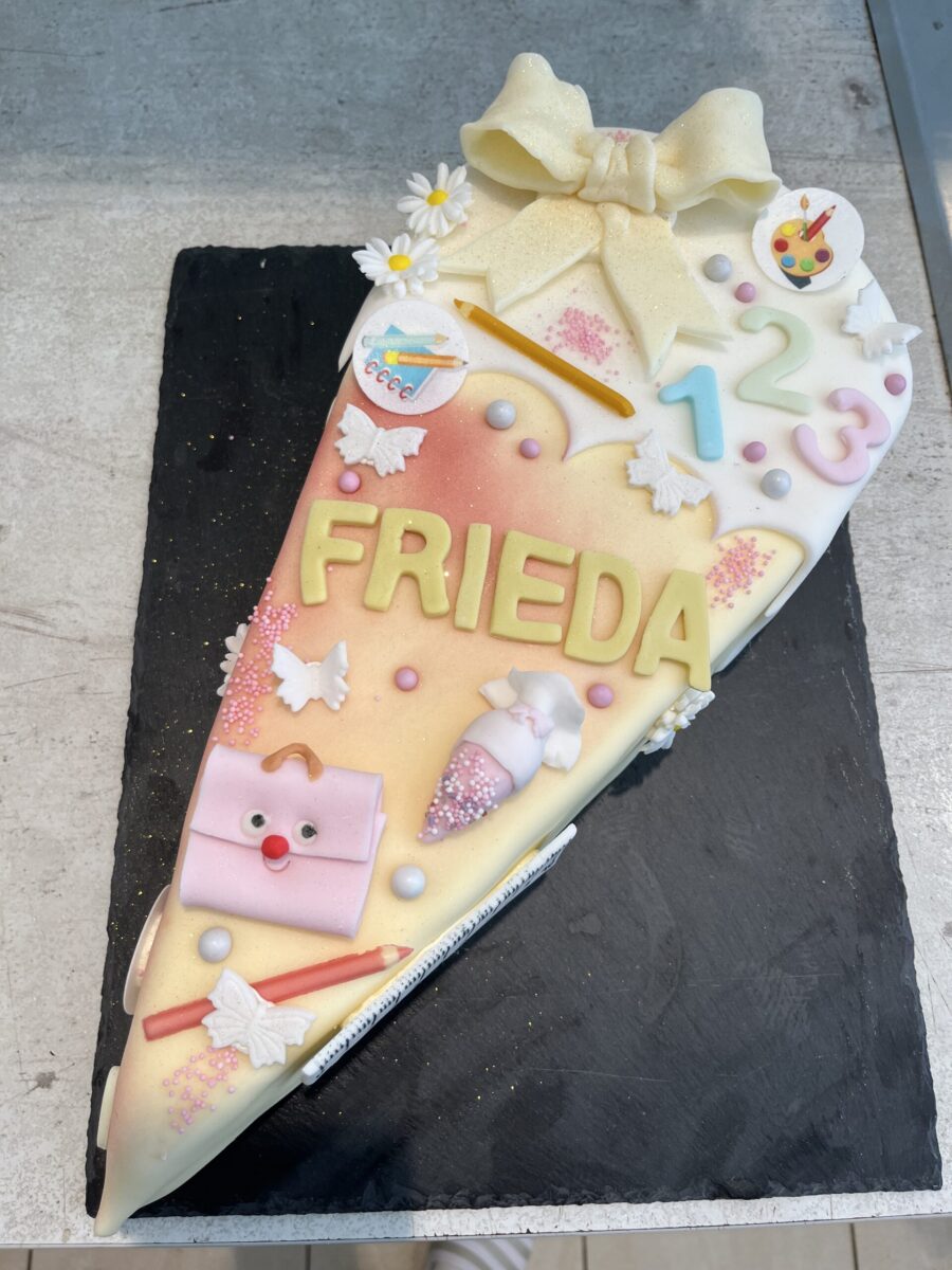 Eine Torte in Form einer Schultüte, verziert mit einer großen gelben Schleife, Gänseblümchen, Zahlen, Bleistiften, Schulsachen und dem Namen "FRIEDA" in gelben Buchstaben. Die Torte hat ein pastellrosa und weißes Design.